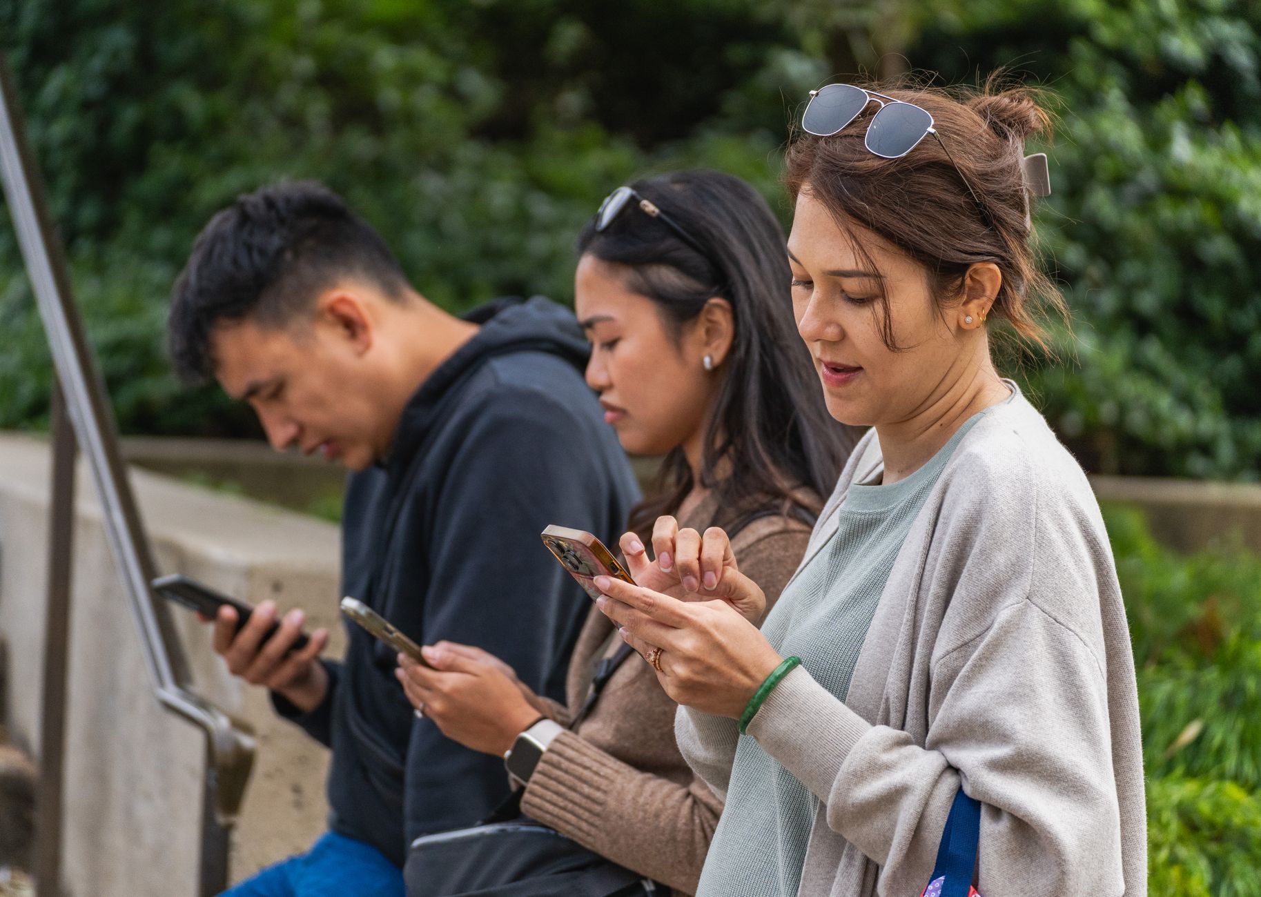 Three young adults focused on their smartphones while sitting outdoors during the day. — How to Choose a New Internet Connect